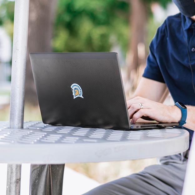 A student wearing a gray baseball cap ponders with his hand to his mouth while they stare at their laptop
