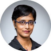A person with short black hair and blue glasses is wearing a black shirt and gold jewelry against a plain backdrop.